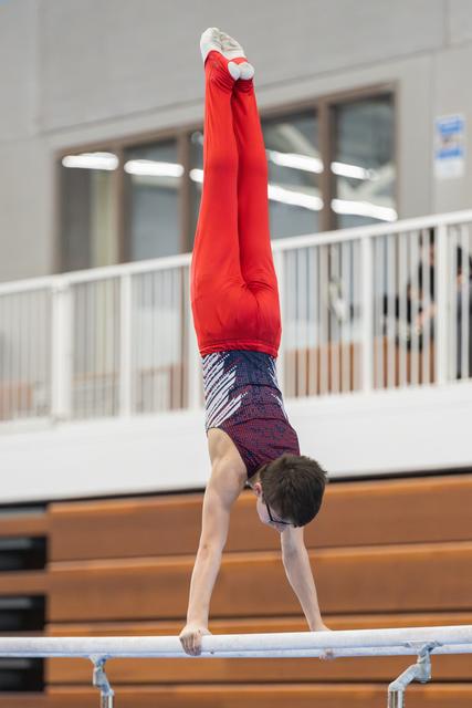 Young gymnast performs vertical handstand on horizontal bar in gymnasium, displaying strength and balance in red outfit