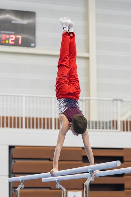 Young gymnast performs handstand on parallel bars wearing red shirt and patterned shorts in indoor gymnasium