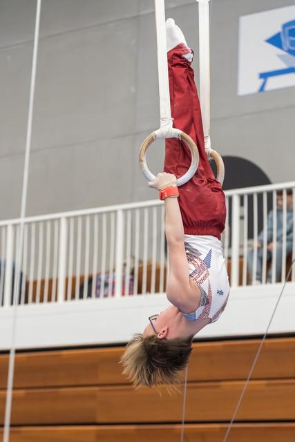 Young gymnast hangs upside down on still rings, demonstrating strength and control in a gym with spectators in background
