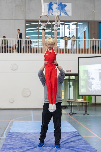 Young gymnast in red uniform hangs from rings while coach supports from below in indoor training facility