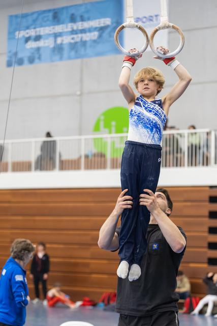 Young gymnast holds rings overhead while being lifted by coach during training session in indoor gymnasium