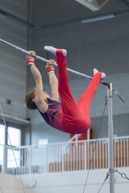 Young gymnast in red outfit performs inverted hang on high bar, legs extended upward in indoor training facility