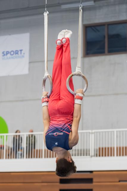 Young gymnast performing an inverted hang on rings, body fully extended upside down in red leotard at indoor facility