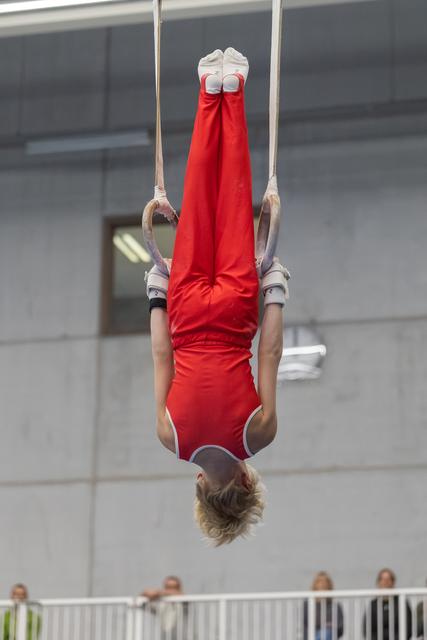 Gymnast performs inverted position on rings, body fully extended vertically with red uniform and white socks