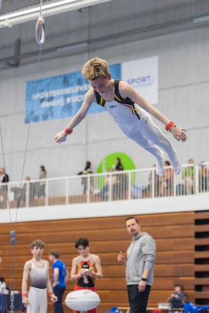 Young gymnast executes airborne trampoline dismount while coach and teammates watch from below in indoor gymnasium