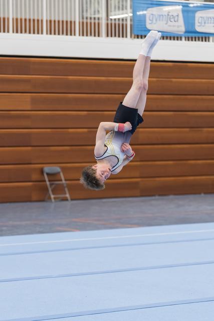 Young gymnast performing an inverted aerial backflip during floor routine in gymnasium with wooden bleachers behind