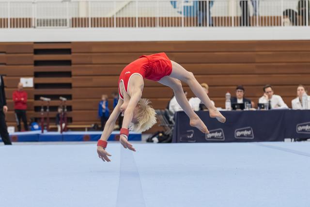 Gymnast performs a backwards somersault during floor routine at indoor gymnastics meet with judges watching