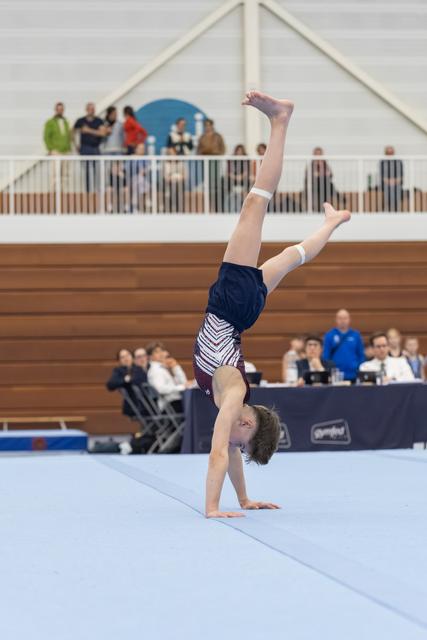 Young gymnast executes a handstand during floor routine, legs extended vertically, in front of judges and spectators