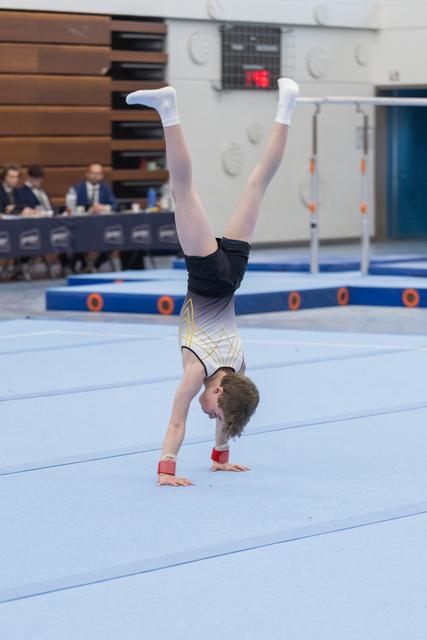 Gymnast executes a vertical handstand on blue floor mat, legs extended straight up, while judges observe from background