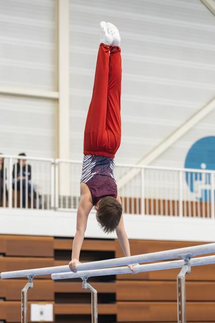 Young gymnast performs a vertical handstand on parallel bars, wearing red top and patterned shorts in indoor training facility