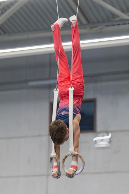 Young gymnast performing an inverted hang on rings, legs extended vertically upward in red uniform during training