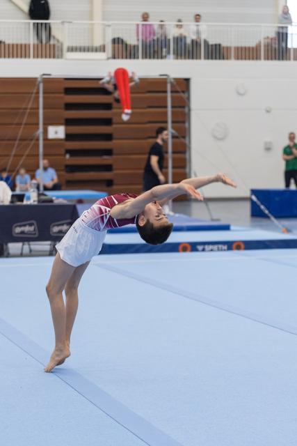 Gymnast executes elegant backward bend during floor routine, demonstrating exceptional flexibility on blue competition mat