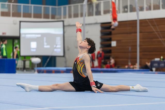 Male gymnast executes a floor split with one arm raised, demonstrating flexibility during his routine in the gymnasium