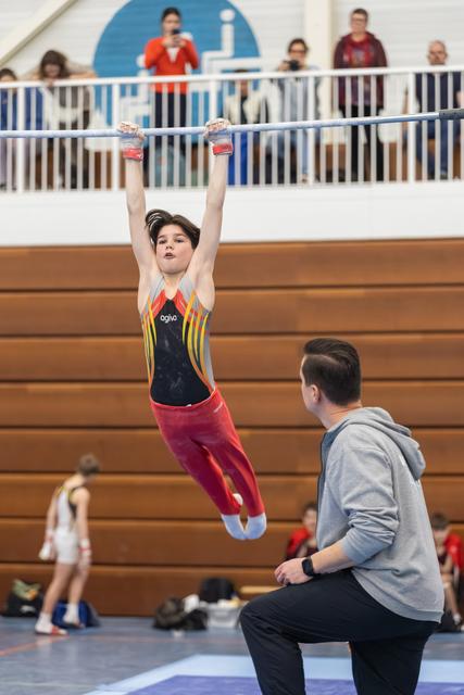 Young gymnast in colorful leotard performs a hanging routine on the bar while coach observes closely below