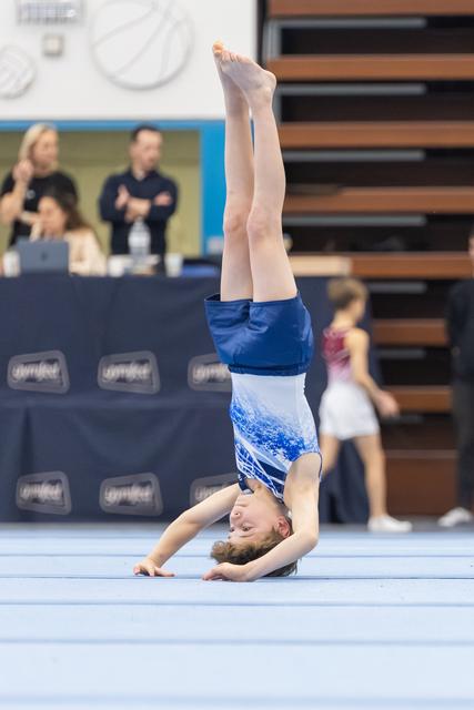 Young gymnast performs a precise headstand on floor mat, legs extended vertically, wearing blue and white leotard