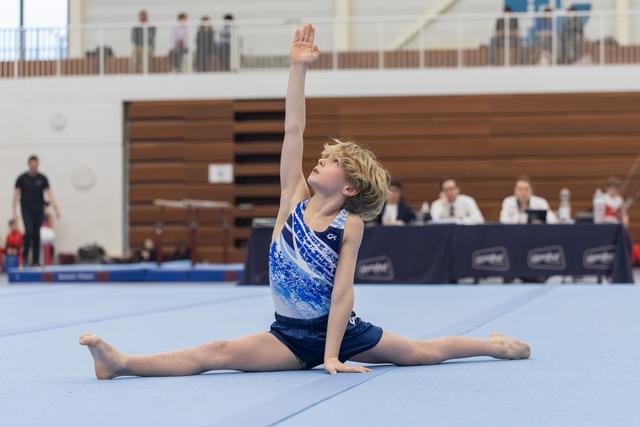 Young gymnast performs a graceful splits pose with raised arm during her floor exercise routine at an indoor gymnastics meet