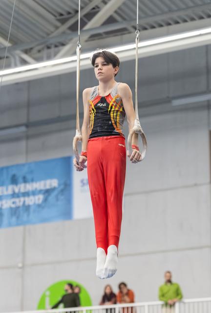 Gymnast performing still rings routine in red pants and flame-design leotard, demonstrating intense focus and control