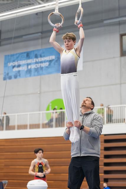 Young gymnast holds onto rings while coach supports him from below during training at indoor gymnasium