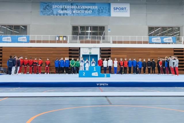 Gymnastics teams lined up behind awards podium in sports hall, with first, second, and third place positions displayed