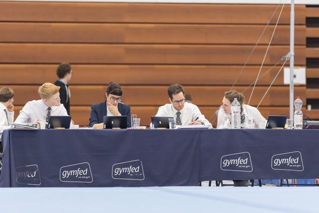 Panel of judges seated at a table with laptops and water bottles, reviewing scores during a gymnastics meet