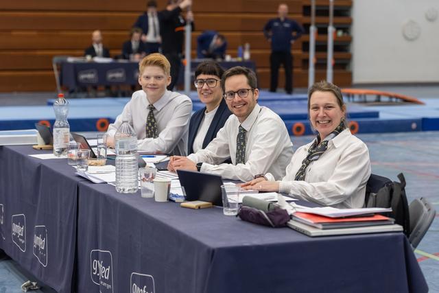 Four judges in white shirts and matching ties sit at a table with water bottles and papers, smiling at the camera