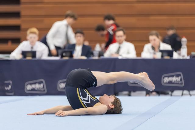 Male gymnast performs a tucked backward roll during floor routine while judges observe from their table in background