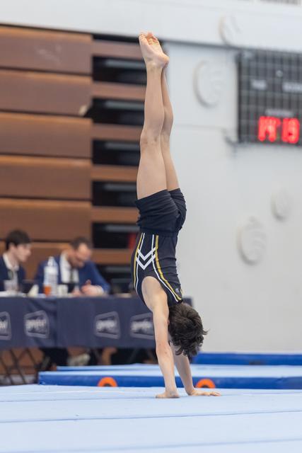 Male gymnast performing a handstand with legs extended upward during routine on blue mat at indoor venue