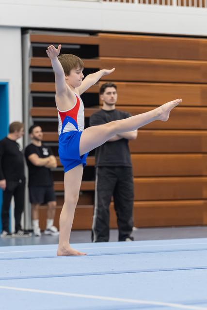 Young male gymnast executes an arabesque pose with pointed toes and extended arms during floor routine on blue mat