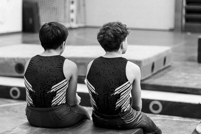 Two young gymnasts in matching black and white leotards sit side by side on the floor, viewed from behind