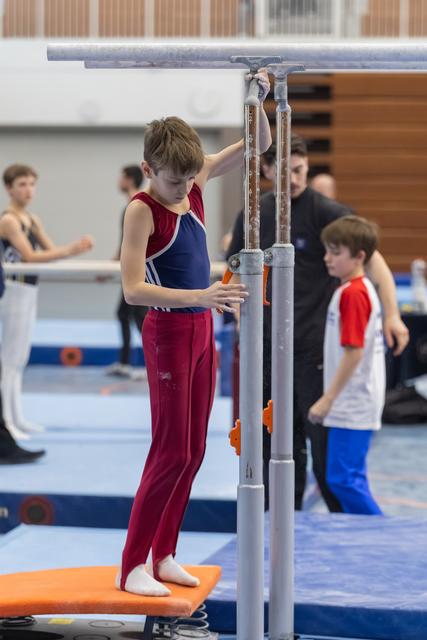 Young gymnast in maroon and blue leotard adjusts parallel bars height while other athletes practice in background