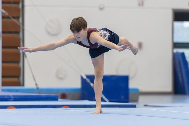 Young gymnast performs arabesque balance on floor exercise mat, arms extended with focused concentration in training facility