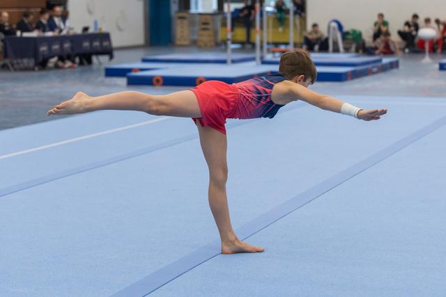 Young gymnast performs arabesque balance on floor mat, arms extended horizontally during routine at indoor gymnastics meet