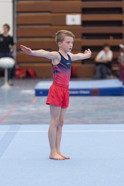 Young male gymnast performs floor routine with arms extended, wearing red gradient leotard in gymnasium