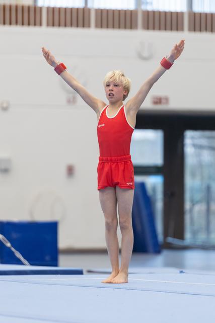 Young gymnast in red leotard stands with arms raised high on floor mat, displaying confident posture in gymnasium