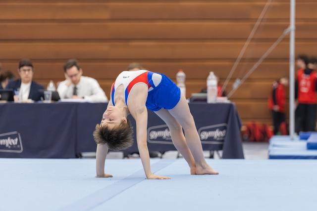 Young gymnast performs a dramatic backbend bridge position on floor mat during routine at indoor gymnastics event