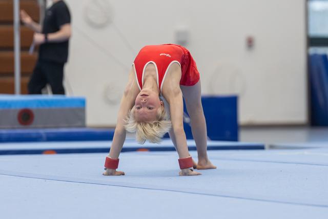 Young blonde gymnast performing a backbend on floor mat, demonstrating flexibility during practice in training facility