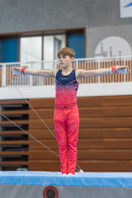 Young gymnast stands on balance beam with arms extended horizontally, wearing pink gradient leotard in training facility