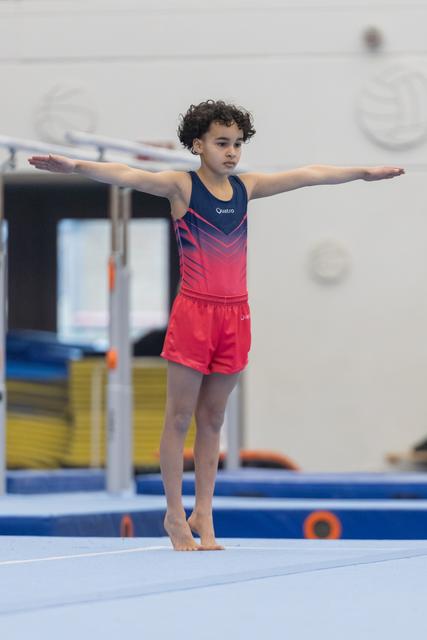 Young gymnast in red and navy leotard performs a T-pose balance on the mat with focused expression during training