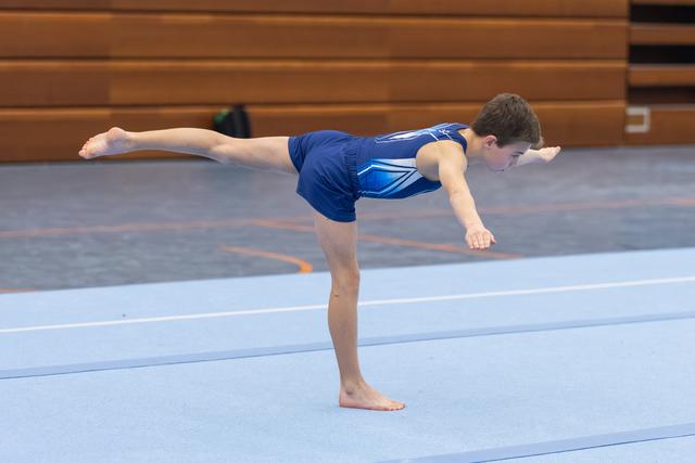 Young gymnast in blue uniform holds an arabesque balance position on floor mat with focused concentration