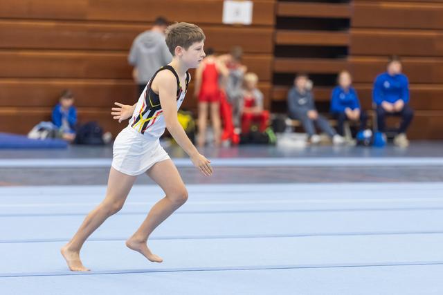 Young gymnast in white leotard running barefoot across blue floor mat during routine, spectators visible in background