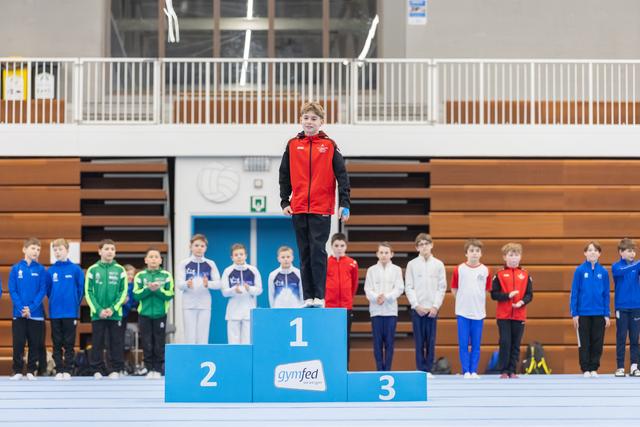 Young gymnast in red vest stands proudly atop first place podium while fellow competitors line up behind at indoor sports hall