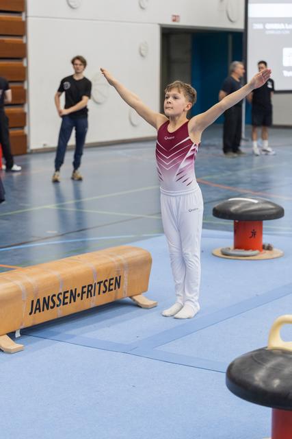 Young gymnast strikes triumphant pose with arms raised after routine, while teammates observe in background
