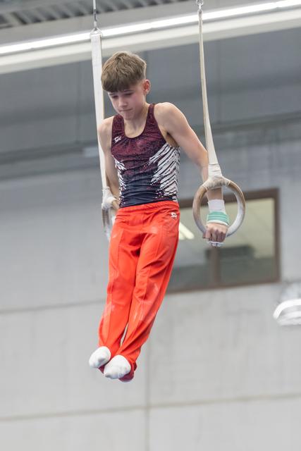 Young male gymnast performs still rings routine with focused expression, wearing orange pants and patterned leotard in training facility