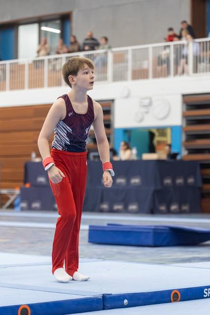 Young gymnast in purple leotard and red pants stands thoughtfully on floor mat, gazing upward during indoor gymnastics event