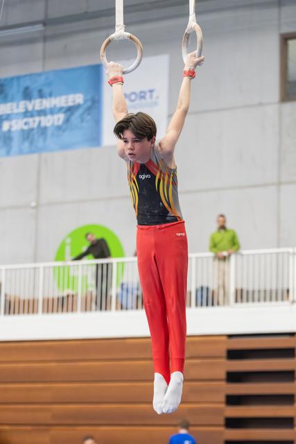 Young gymnast in red pants and striped leotard hangs from rings with arms extended, showing focused concentration