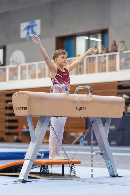 Young male gymnast dismounting pommel horse with arms raised triumphantly, wearing maroon and white leotard in training facility