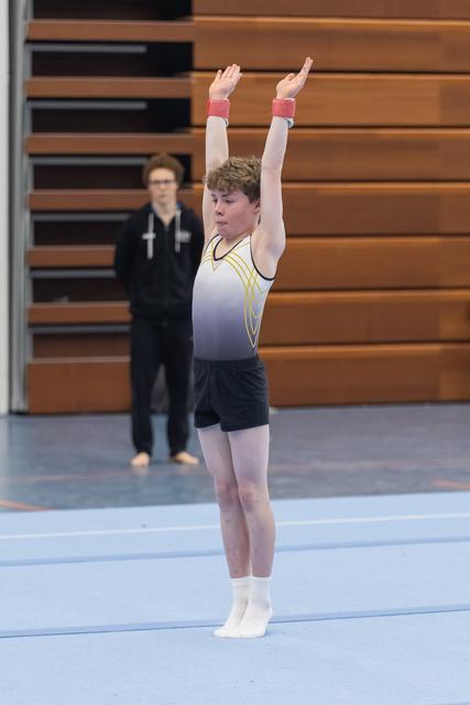 Young gymnast completes floor routine with arms raised, displaying focused determination as judge observes in background