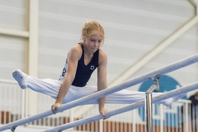 Young blonde gymnast performs on parallel bars with focused concentration, legs extended horizontally in white and black uniform