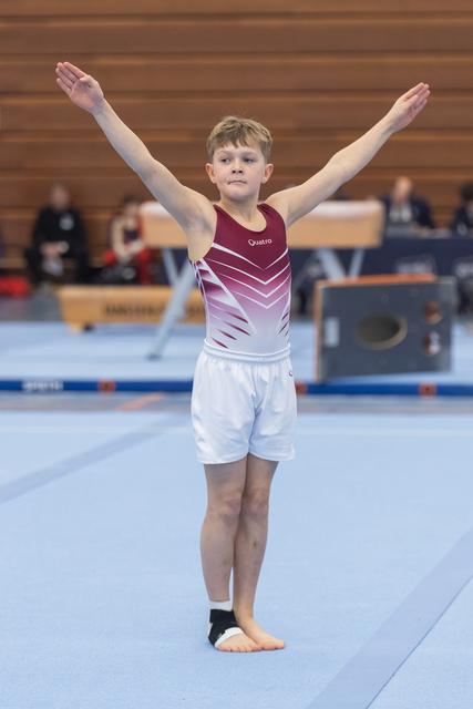 Young boy in maroon and white leotard strikes finishing pose with arms raised on gymnastics floor mat