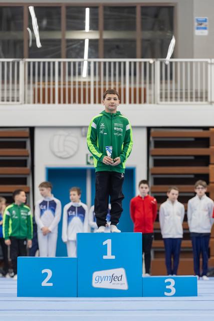 Young gymnast in green jacket stands proudly on first place podium holding medal, with competitors lined up behind